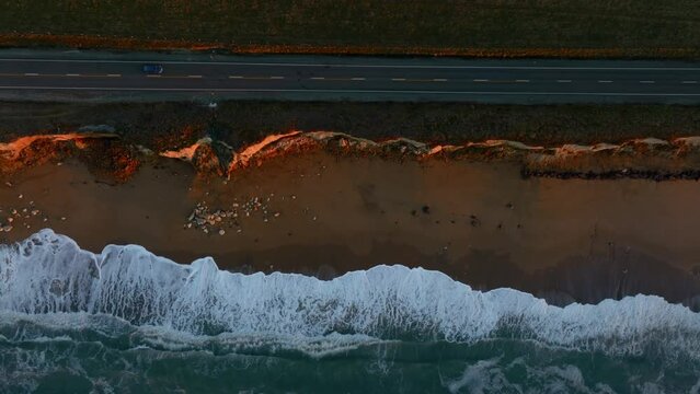 Aerial: Ocean beach surf, country road and  farmland in sunrise. Oamaru, Otago, New Zealand.