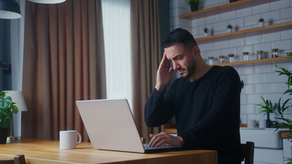 adult man having pain from a headache or migraine rubbing his head while using laptop in the kitchen at home