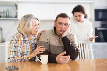 Family members quarreling in the kitchen and old woman trying to calm angry man sitting at the table