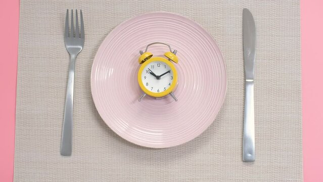 Empty plate with alarm clock and female hand on pink background, intermittent fasting concept.