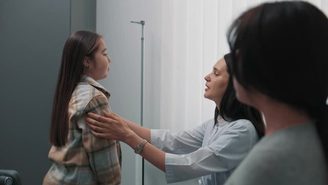 Female endocrinologist checks thyroid gland of Asian girl in hospital. Young patient undergoes examination or does checkup in clinic. Mother talks with medical professional. Concept of healthcare.