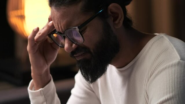 Sad and stressed Indian man with a headache, depression, exhaustion from work, fatigue from overwork sits on the sofa in the living room in the evening at home.