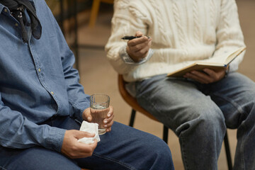 Closeup of unrecognizable person holding tissue and glass of water during therapy session with psychologist speaking in background, copy space