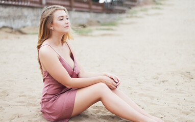 Young woman in pink dress sitting on sand and enjoys sea.