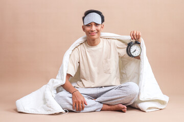 Young Asian guy wear sleep mask, sitting on floor covered by blanket and holding an alarm clock