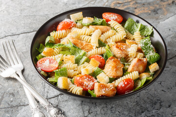 Chicken Caesar Salad with Pasta fusilli, romaine lettuce, parmesan, tomatoes and croutons close-up in a bowl on the table. Horizontal 