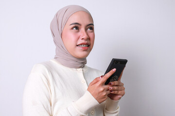 Young muslim girl with hijab and braces teeth holding smartphone while looking up, think of something