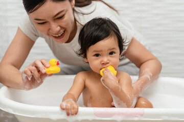 mother bathing her cheerful infant baby with foam bubbles and playing rubber duck in bathtub