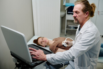 Obraz premium Mature endocrinologist with ultrasonic probe sitting in front of ultrasound machine in medical office and looking at screen with sonogram