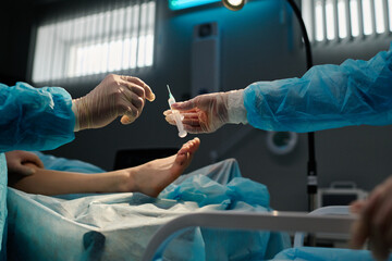 Gloved hand of surgeon taking syringe with local anaesthesia during surgical operation against leg...