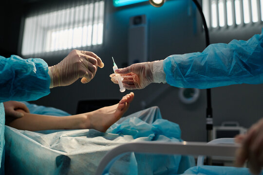 Gloved hand of surgeon taking syringe with local anaesthesia during surgical operation against leg of patient lying on operating table