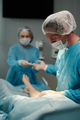 Mature surgeon taking medical tool from hand of female assistant in medical scrubs, gloves, cap and mask during operation