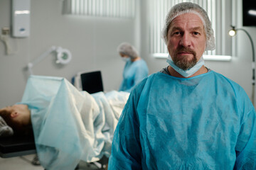 Serious male surgeon in medical scrubs and protective cap looking at camera against operating table with patient covered with napkin