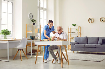 Senior man doing puzzles while sitting at desk in cozy retirement home interior. Friendly nurse helping demented old man while playing games and doing puzzles. Senior care, Alzheimer disease concept