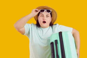 Deurstickers Portrait of shocked surprised fat woman with open mouth in straw hat holding suitcase raising sunglasses on studio yellow background. Funny tourist is going on summer holiday trip. Vacation concept. © Studio Romantic
