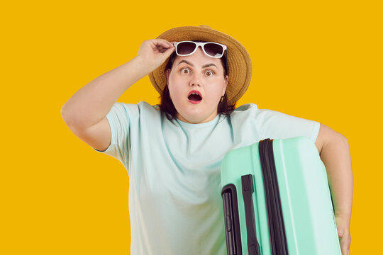 Portrait of shocked surprised fat woman with open mouth in straw hat holding suitcase raising sunglasses on studio yellow background. Funny tourist is going on summer holiday trip. Vacation concept.
