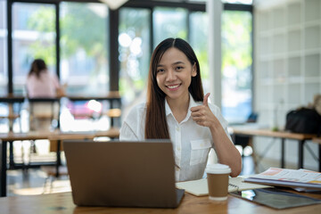 Cheerful young Asian businesswoman giving a thumbs up while working with a laptop at a wooden table in a bright office..