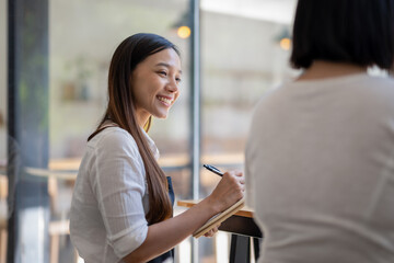 Fototapeta premium A cheerful Asian cafe owner takes an order from a customer, writing on a notepad with a warm and welcoming smile..