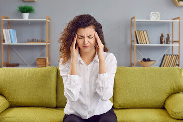 Portrait of a young curly redhead woman suffering from headache disease sitting on the sofa at...