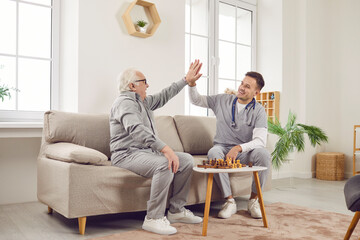 Young nurse man giving high five to a gray-haired senior elderly person while playing chess game...