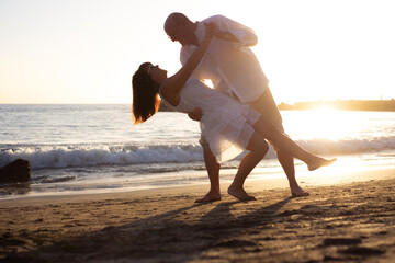 Happy bonding senior couple dancing at the beach enjoying vacation and retirement, two elderly people expressing love and tenderness at sunset - obrazy, fototapety, plakaty