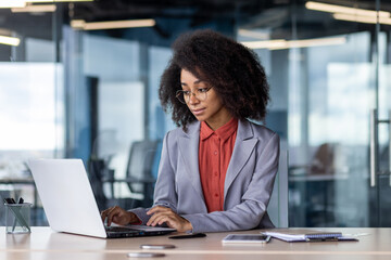 Confident young businesswoman with curly hair working intently on her laptop in a stylish office environment.