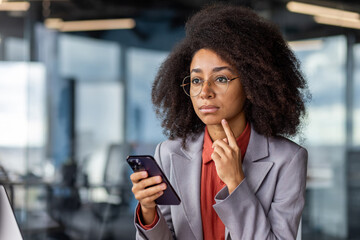 Focused female entrepreneur with curly hair and glasses contemplating while holding a phone in a...