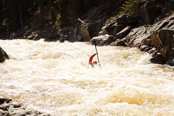 Man in a red kayak paddles through a rough river with large rocks on the shore and in the water