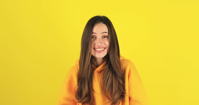 Shocked smiling shy charming confused young woman in yellow hoody posing isolated on yellow background studio. Girl looking at camera smile cover mouth with hand say oops.