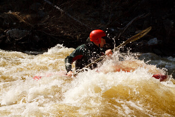 Man in a red helmet kayaking in a river with white water and rocks in the background.