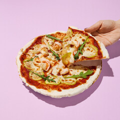 Hand taking a slice of gluten-free pizza with shrimp and zucchini on a pink background