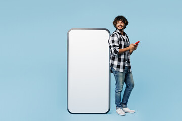 Full body side view young Indian man wears shirt white t-shirt casual clothes big huge blank screen area mobile cell phone using smartphone isolated on plain blue cyan background. Lifestyle concept.