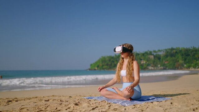 Woman in VR headset meditates on sandy beach, virtual reality transport to exotic locale for mindfulness. Tech offers serene escape, balance, calm. Female practices yoga poses by sea, tranquility.