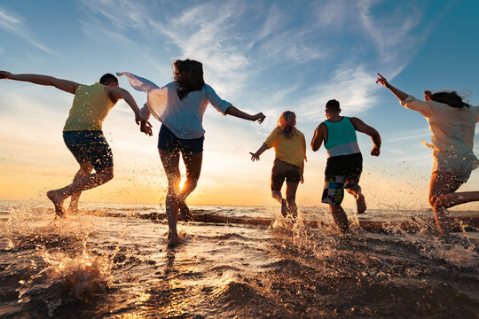 Group of happy young friends are having fun, runs and jumps at sunset beach with lots of splashes