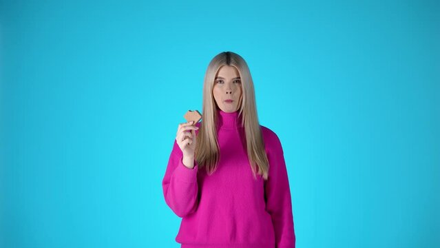 Młoda blond kobieta w r&oacute;żowym swetrze typu golf jedząca kwadratowy batonik i uśmiechająca się z zadowoleniem &mdash; błękitne tło, smak, słodycz i kobiecy styl w slow motion 4K