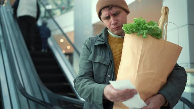 Price rise concept. Surprised male customer holding paper bag with food products looks at paper receipt after shopping in supermarket.