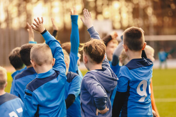 Children's sports team standing in a circle and rising hands up. Kids play sports match. School boys in blue jersey shirts huddling in a team. School sports team compete in a game - obrazy, fototapety, plakaty