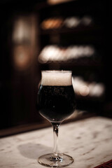 Luscious dark beer in an elegant glass, standing tall on the bar counter. Warm ambiance, ideal for promoting relaxed moments and flavorful experiences.