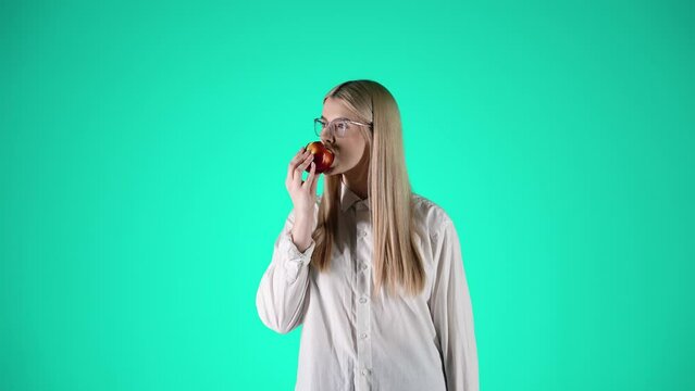 Młoda blond kobieta w białej koszuli i okularach jedząca czerwone jabłko i patrząca w kamerę &mdash; centralne ujęcie na zielonym tle, naturalność i smak w slow motion 4K