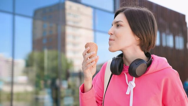 Calm stylish woman with wireless headset around neck eating ice cream in waffle cone and looking aside outdoors. Carefree female having refreshing snack while waiting for fiends no sunny day.