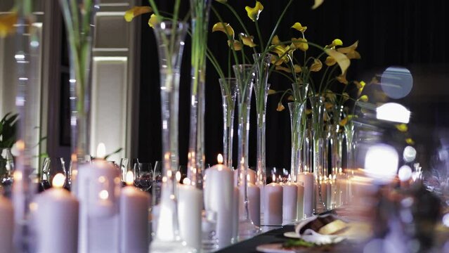 Elegantly arranged table setting for event or celebration. Multiple tall glass vases containing yellow flowers displayed on wooden desk. White candles vary in height enhancing charming atmosphere.