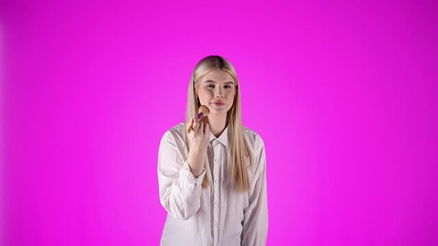 Młoda kobieta w białej koszuli z blond włosami pudrująca pędzlem policzki, brodę i czoło na r&oacute;żowym tle &mdash; beauty, styl i kobieca pielęgnacja w slow motion 4K