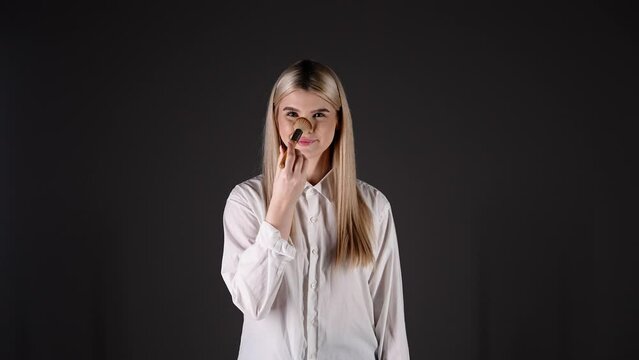 Modelka na czarnym tle uśmiecha się i pudruje twarz oraz nos pędzlem do makijażu. Eleganckie ujęcie beauty pokazujące kobiecy rytuał pielęgnacyjny w stylizowanym portrecie studyjnym