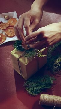 Vertical. A hand tying a ribbon of twine on a gift box wrapped in recycled paper and decorated by dried orange slice and pine leaves. Sustainable zero waste lifestyle