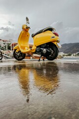 a yellow moped stands on wet asphalt against the backdrop of mountains in cloudy weather