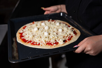 Homemade Pizza Ready for Oven Baking. A person in a black shirt prepares to bake a homemade pizza topped with cheese and tomato sauce on a baking tray.