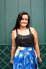 Young beautiful woman with black hair, in black clothes with blue skirt, standing against green door.