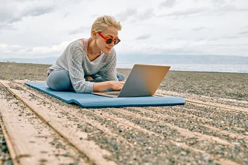 Abwaschbare Fototapete Positive well-fit blond woman in sportswear, seated in lotus pose, using laptop at the beach. Freelance working, wellbeing concept. © Caterina Trimarchi