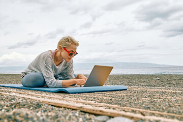 Positive well-fit blond woman in sportswear, seated in lotus pose, using laptop at the beach....