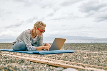  Positive well-fit blond woman in sportswear, seated in lotus pose, using laptop at the beach. Freelance working, wellbeing concept. © Caterina Trimarchi